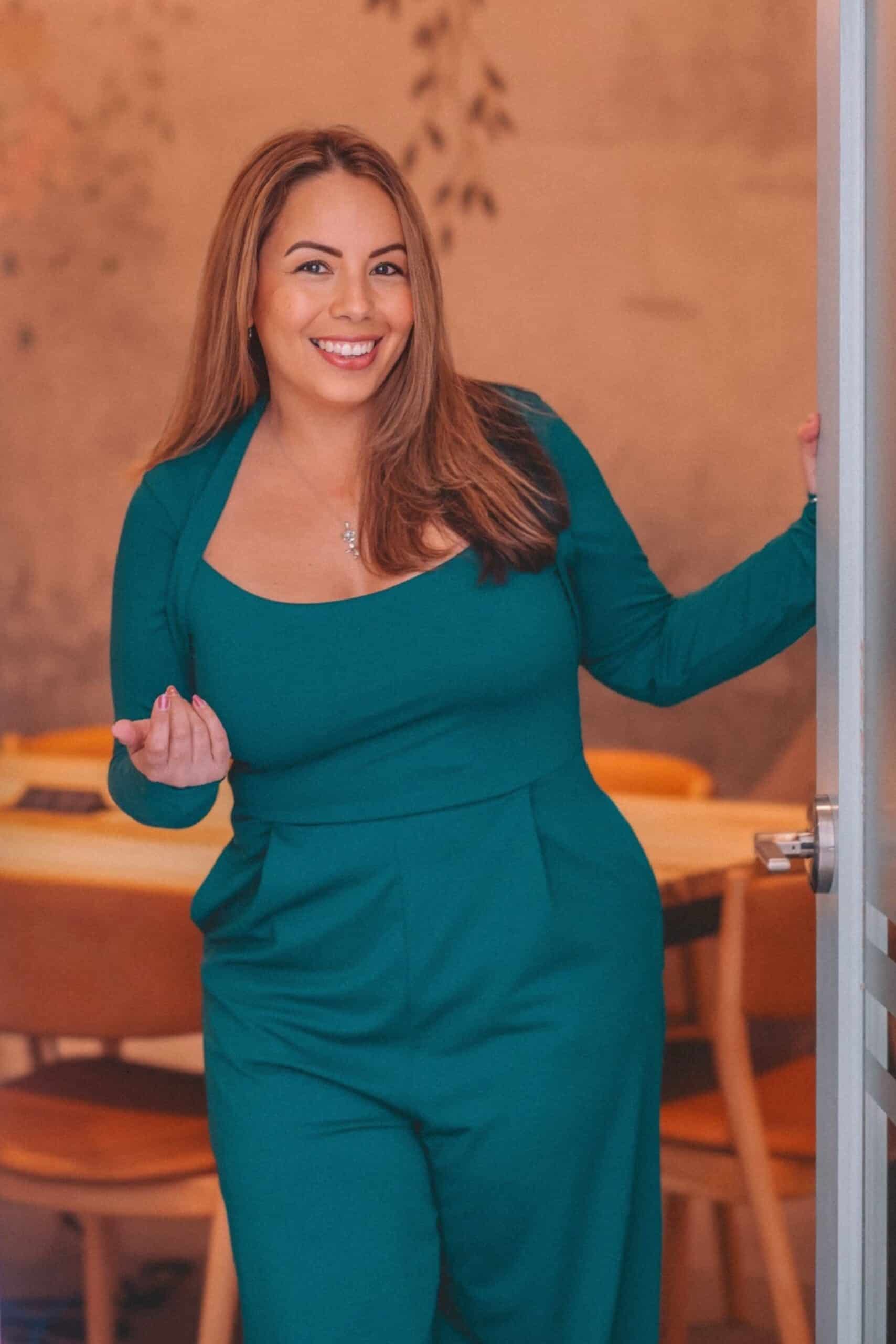A woman with long brown hair, wearing a teal jumpsuit, stands in a doorway smiling and gesturing with her left hand. The room behind her has wooden chairs and a warm, softly lit ambiance.