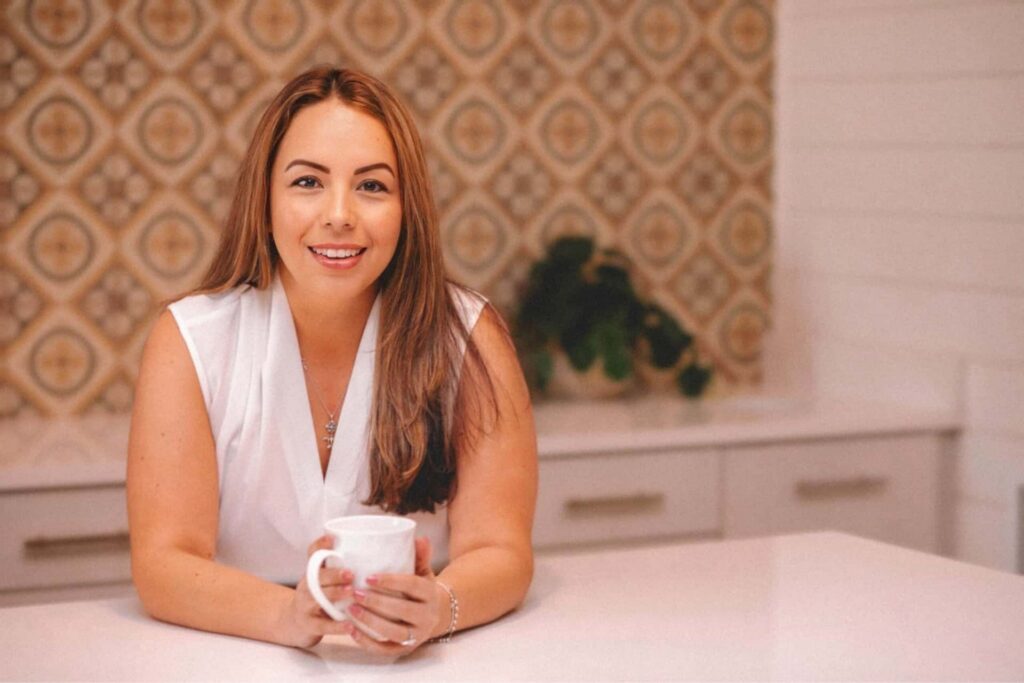 A woman with long brown hair and a white sleeveless top sits at a kitchen counter, smiling and holding a white mug. The background features patterned tiles and white cabinets with a small plant.