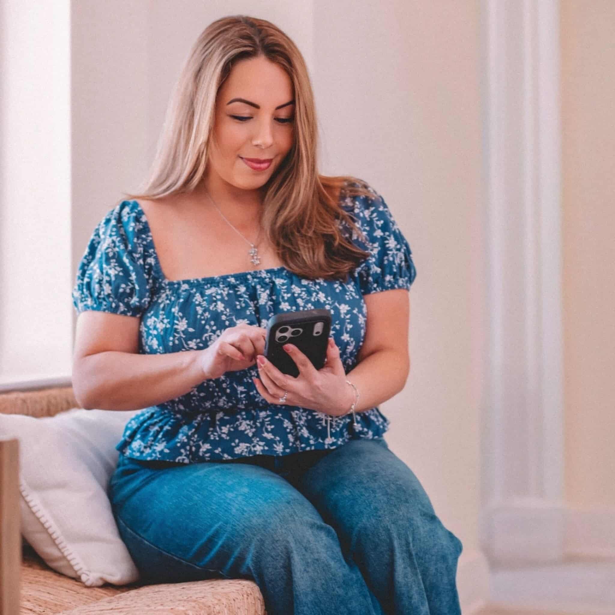 A woman with long brown hair, wearing a blue floral top and jeans, sits on a woven chair indoors and looks at her smartphone, smiling slightly.