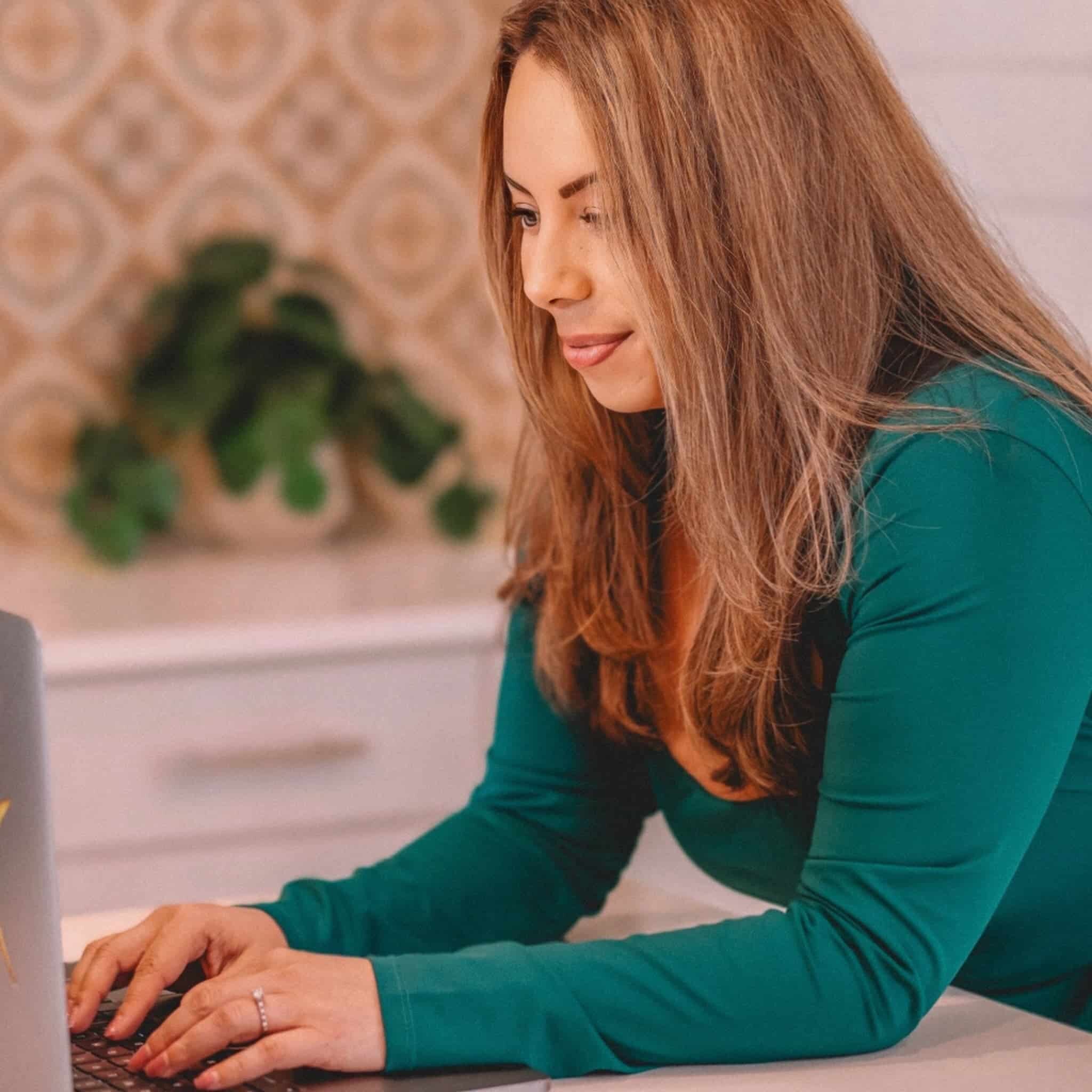 A woman with long light brown hair, wearing a green top, is typing on a laptop at a desk. The background features a blurred plant and patterned wall.