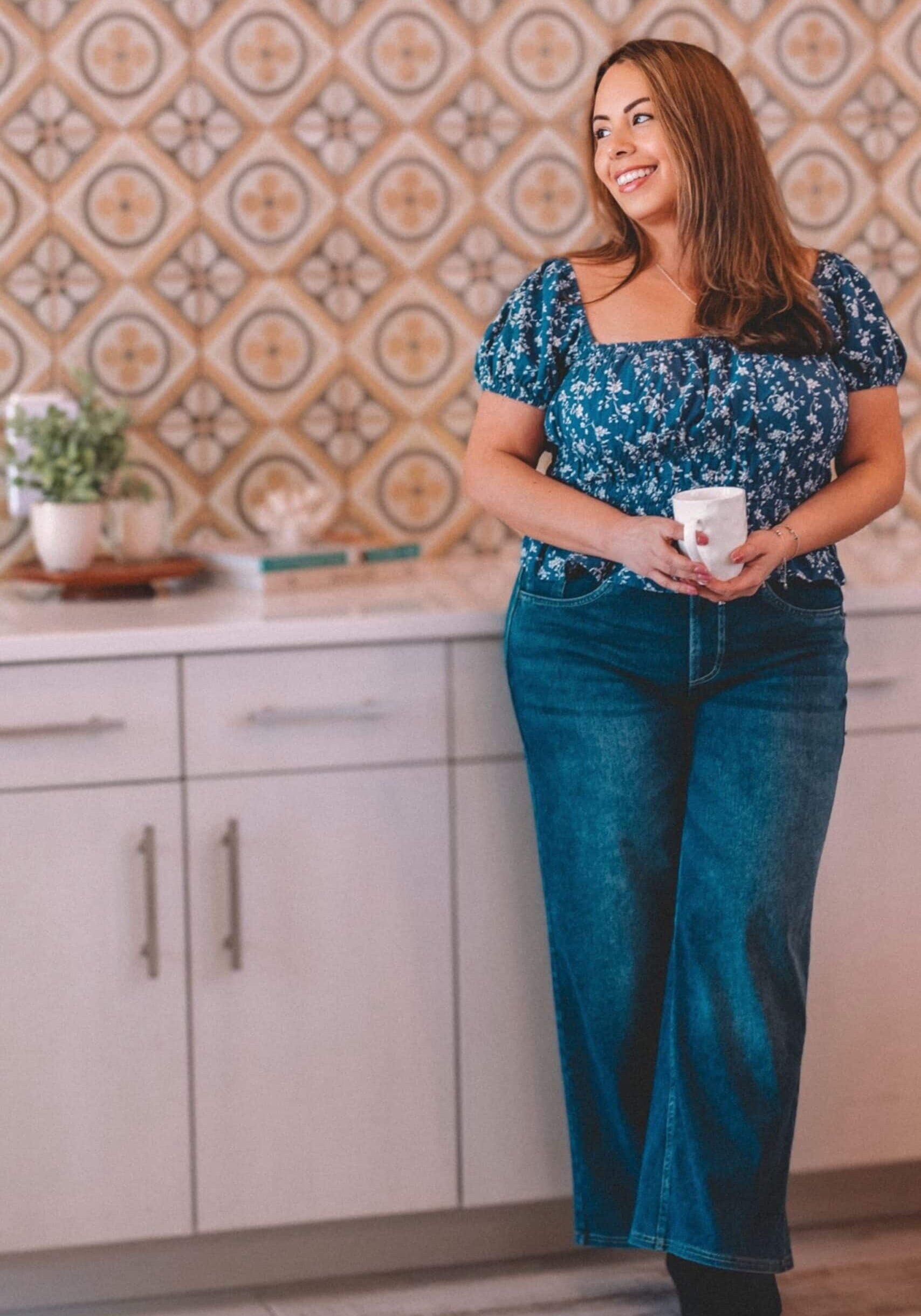 A woman with long brown hair wearing a blue floral top and jeans stands in a kitchen, smiling and holding a mug. The background features patterned wallpaper and white cabinets with a plant on the counter.